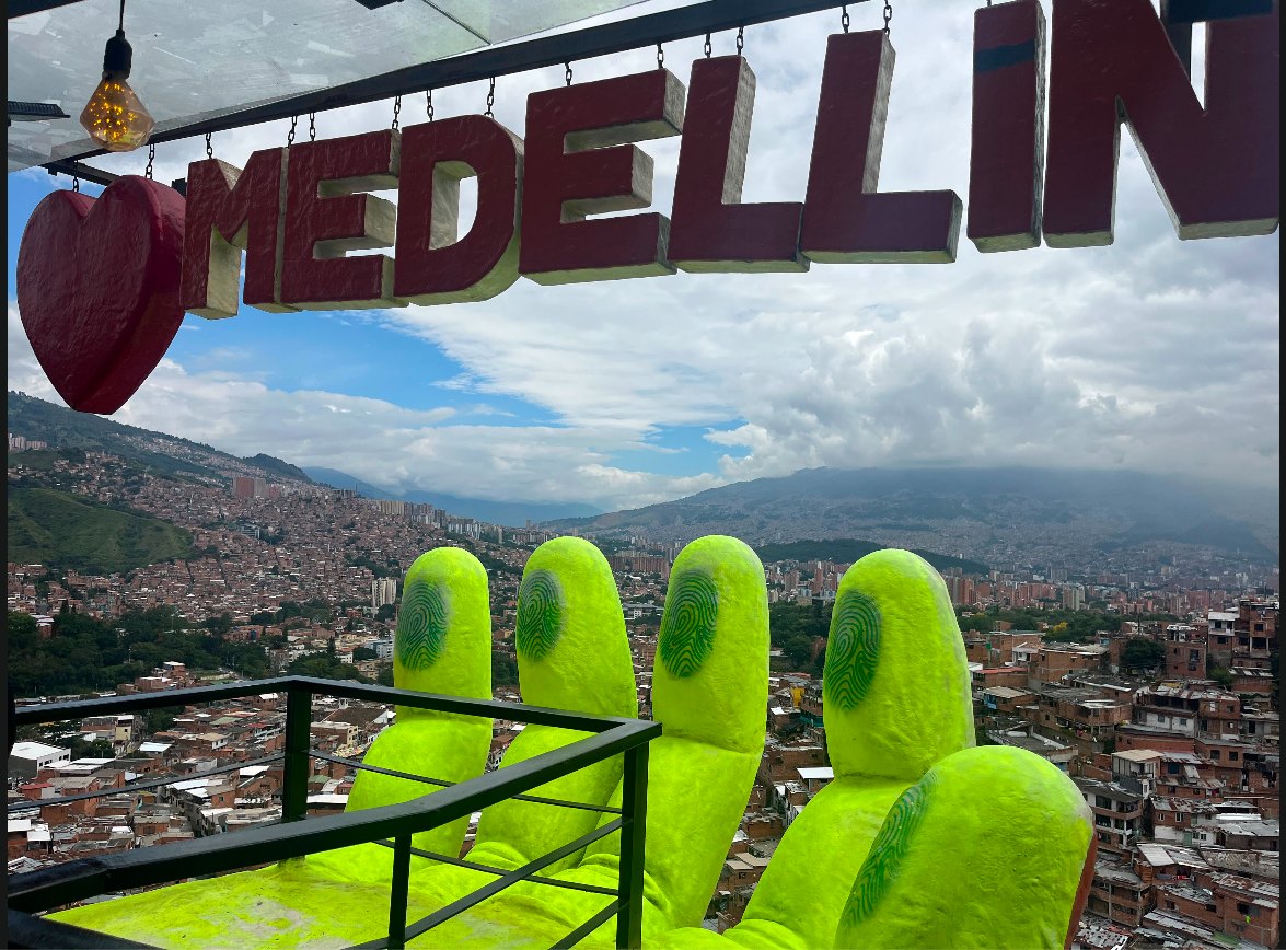 The giant neon-green Monumento a la Vida hands sculpture beneath a hanging Medellin sign, overlooking the hillside barrios and Aburra Valley skyline of Medellin, Colombia